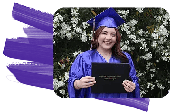 SIATech student wearing graduation cap and gown holding up diploma in front of wall of flowers